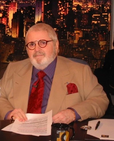 Jô Soares seated at a table in a beige suit and red tie, holding papers during an interview or studio appearance.